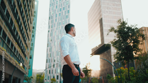 Mature businessman with neat beard wearing white shirt stands in the financial district in the city. Successful man looks around on modern buildings background