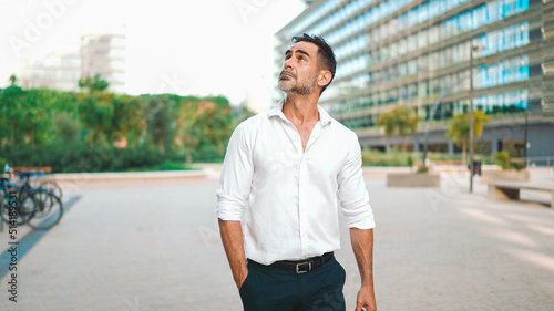 Mature businessman with neat beard wearing white shirt on his way to the office in the financial district in the city. Successful man Looks at the upper floors of modern buildings