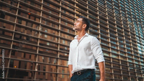 Mature businessman with neat beard wearing white shirt on his way to the office in the financial district in the city. Successful man Looks at the upper floors of modern buildings