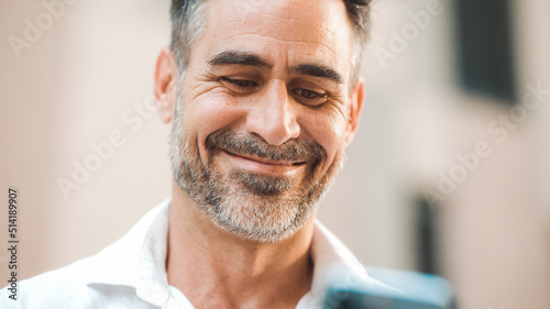 Mature businessman with neat beard uses mobile phone sits on bench in the financial district in the city. Successful man scrolls through information on smartphone and smiles