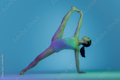 Portrait of young sportive girl doing stretching exercises isolated over blue studio background in neon light. Twine position
