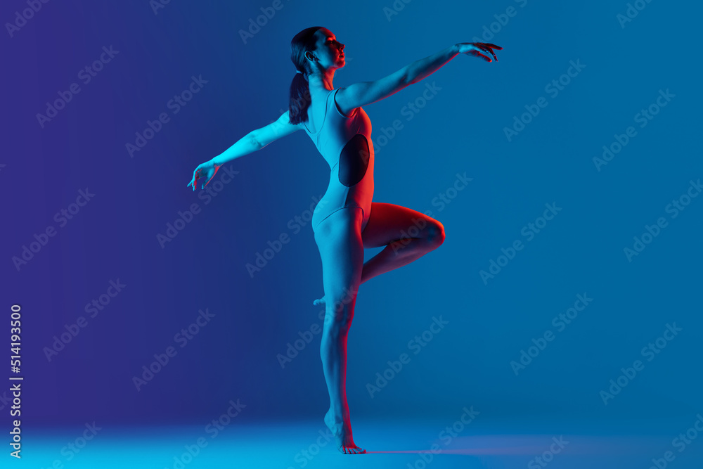 Portrait of young sportive girl doing stretching exercises isolated over blue studio background in neon light. Dancing moves