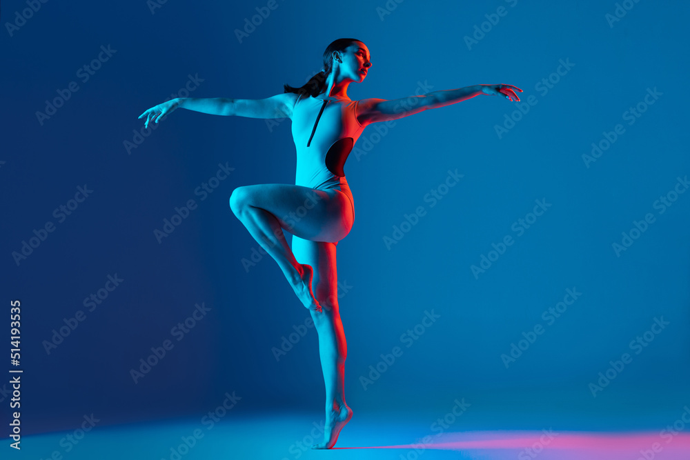 Portrait of young sportive girl doing stretching exercises, training, dancing isolated over blue studio background in neon light