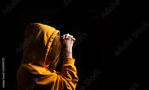 Unrecognizable woman with a hood put on her head praying on a black background. Side view.