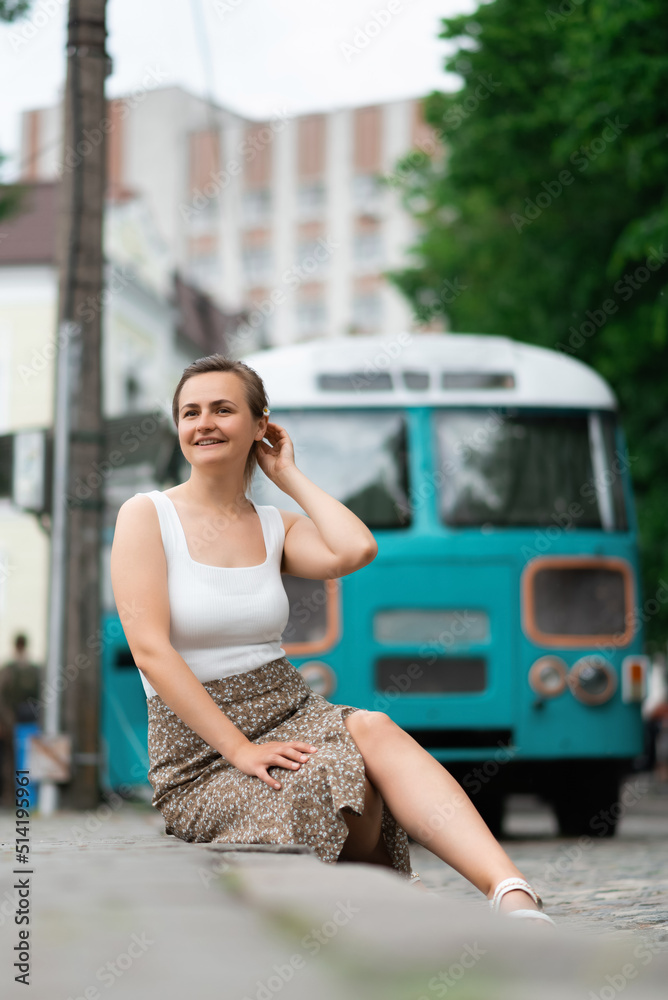An attractive young woman in a white blouse and brown skirt is sitting on a sidewalk in the background of an old school bus