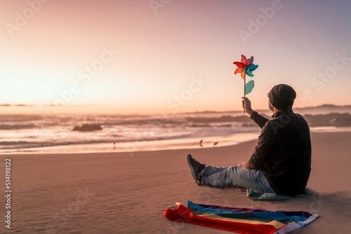 silhouette of a person sitting on the beach outdoors with rainbow flag holding windmill toy