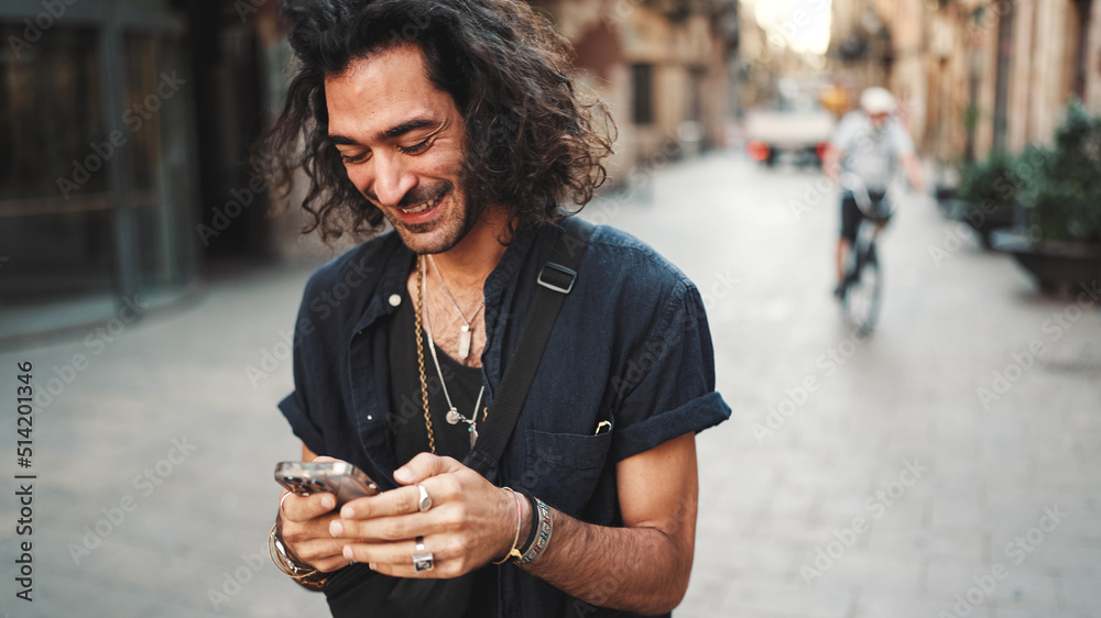 Young attractive italian guy with long curly hair and stubble is using mobile phone at old buildings background. Stylish man with an earring in his ear and lot of chains writing message on smartphone