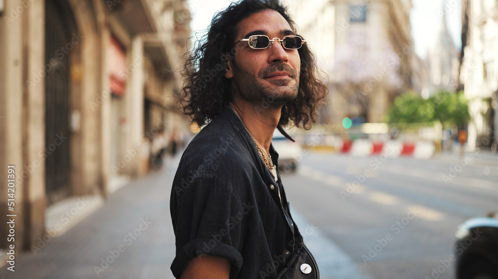 Young italian guy with long curly hair and stubble walks smiling down the street. Stylish man with sunglasses and lot of chains smiling and looking around