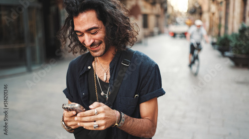 Young attractive italian guy with long curly hair and stubble is using mobile phone at old buildings background. Stylish man with an earring in his ear and lot of chains writing message on smartphone