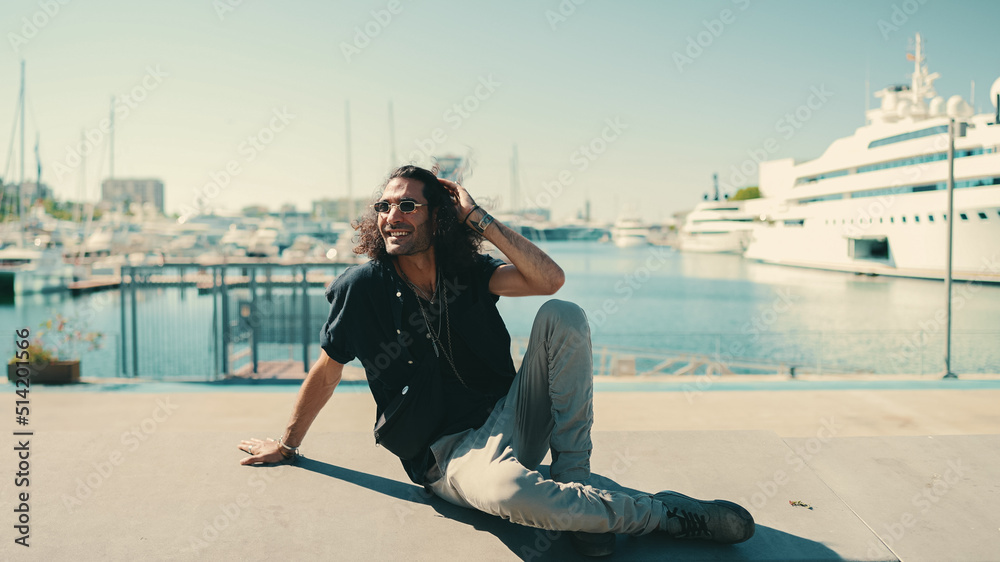 Young italian guy with long curly hair and stubble sits in harbor and looks at ships and yachts