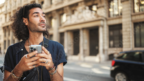Young attractive italian guy with long curly hair and stubble is using mobile phone. Stylish man with writes message on smartphone