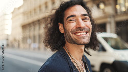 Young italian guy with long curly hair and stubble stands on the sidewalk next to the road, looks around and smiles