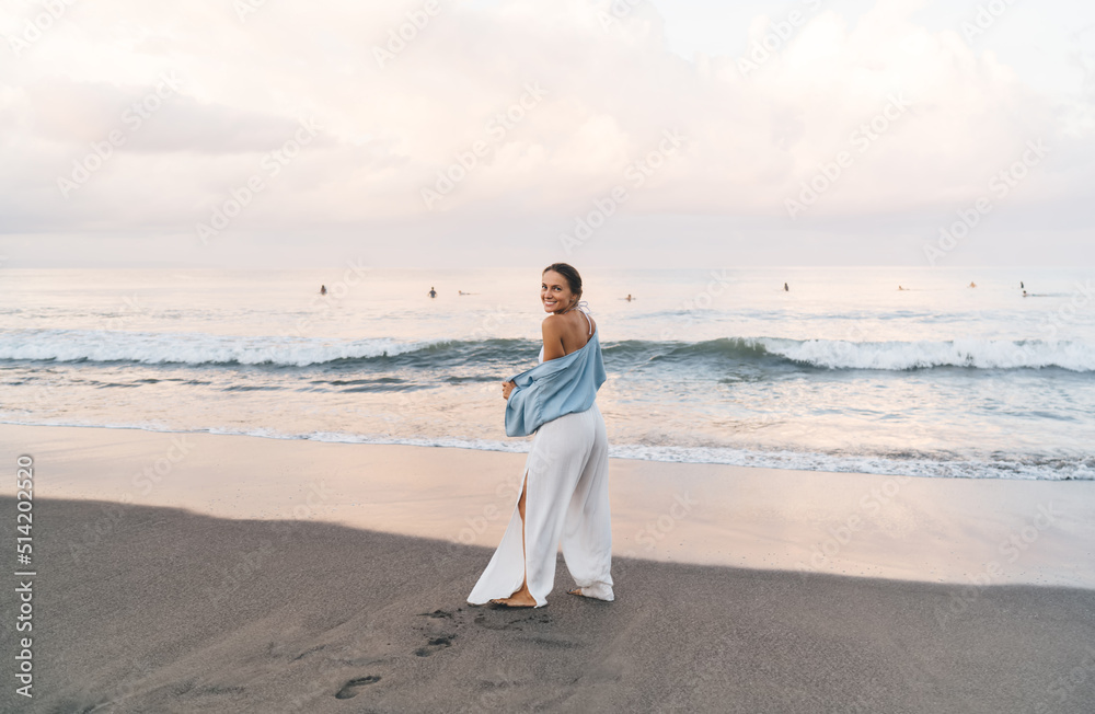 Gorgeous Caucasian woman enjoying vacations while walking on the beach feeling calm and carefree. Beautiful healthy female in white summer cloth strolls on seashore during holidays travel Gorgeous Caucasian woman enjoying vacations while walking on the beach feeling calm and carefree. Beautiful healthy female in white summer cloth strolls on seashore during holidays travel