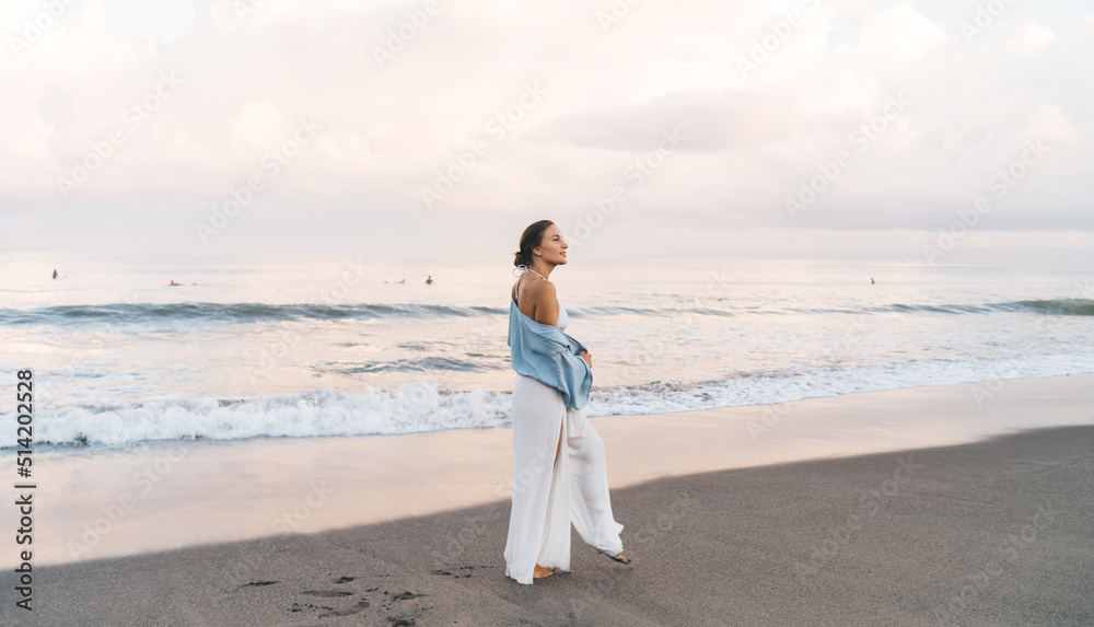 Gorgeous Caucasian woman enjoying vacations while walking on the beach feeling calm and carefree. Beautiful healthy female in white summer cloth strolls on seashore during holidays travel Gorgeous Caucasian woman enjoying vacations while walking on the beach feeling calm and carefree. Beautiful healthy female in white summer cloth strolls on seashore during holidays travel