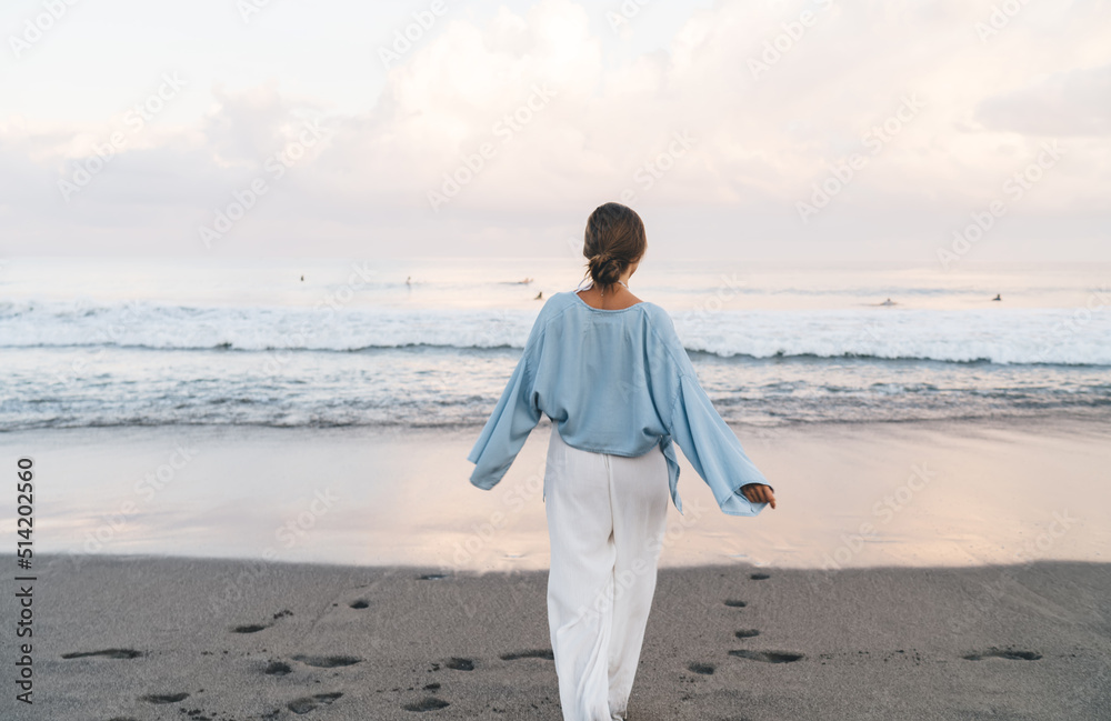 Gorgeous Caucasian woman enjoying vacations while walking on the beach feeling calm and carefree. Beautiful healthy female in white summer cloth strolls on seashore during holidays travel