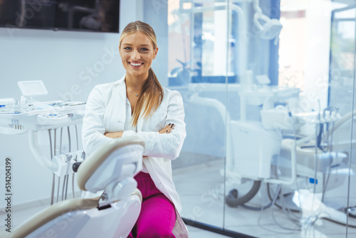 Portrait of smiling female dentist in scrubs standing with arms crossed at clinic. Portrait of a Caucasian woman dentist, sitting in her office next to a dentist chair, smiling