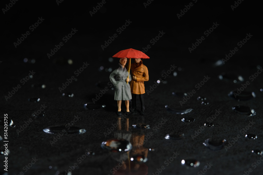 Miniature people toy figure photography. A couple using umbrella, walking on the floor full of droplet. Dark cloudy sky background