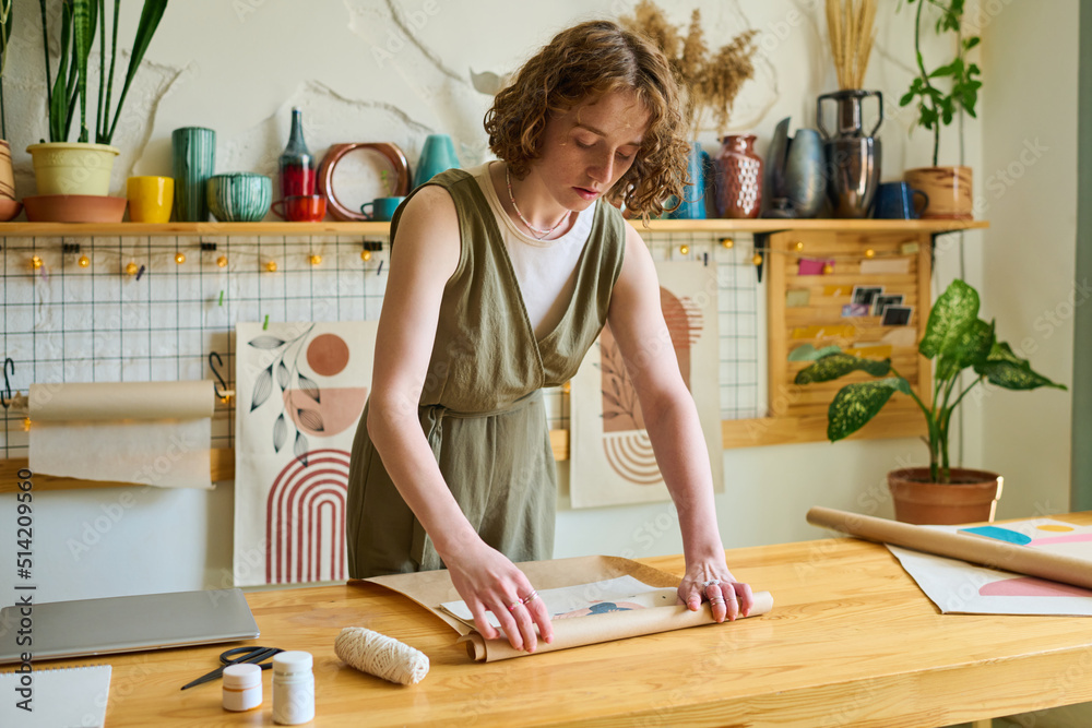 Young female clerk bending over table while wrapping self made abstract painting into paper while packing order of client in workshop Young female clerk bending over table while wrapping self made abstract painting into paper while packing order of client in workshop