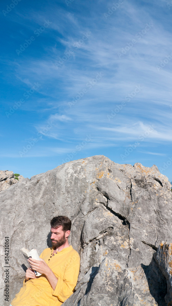 Hombre blanco barbudo leyendo un libro entre rocas en playa rocosa Hombre blanco barbudo leyendo un libro entre rocas en playa rocosa