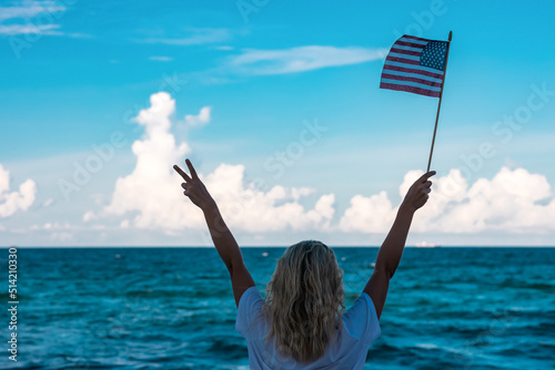 A blonde girl is standing on the ocean. In his right hand he holds an American flag, and his left hand shows 2 fingers up. Soft focus