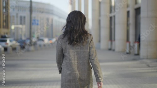 View from the back: a young woman with dark hair in a coat walks forward. Close to office buildings. Slow motion 4k footage