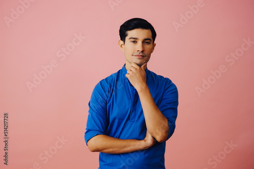 Portrait hispanic latino man with arms crossed hand on chin black hair smiling handsome young adult blue shirt over pink background looking at camera studio shot