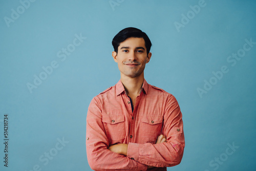 Portrait smiling handsome young adult latino man with arms crossed and hand on chin black hair and pink shirt over blue background looking at camera studio shot