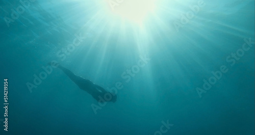 Beautiful underwater image freediver woman silhouette swimming from surface with sun rays to depth of blue sea. Unfocused image