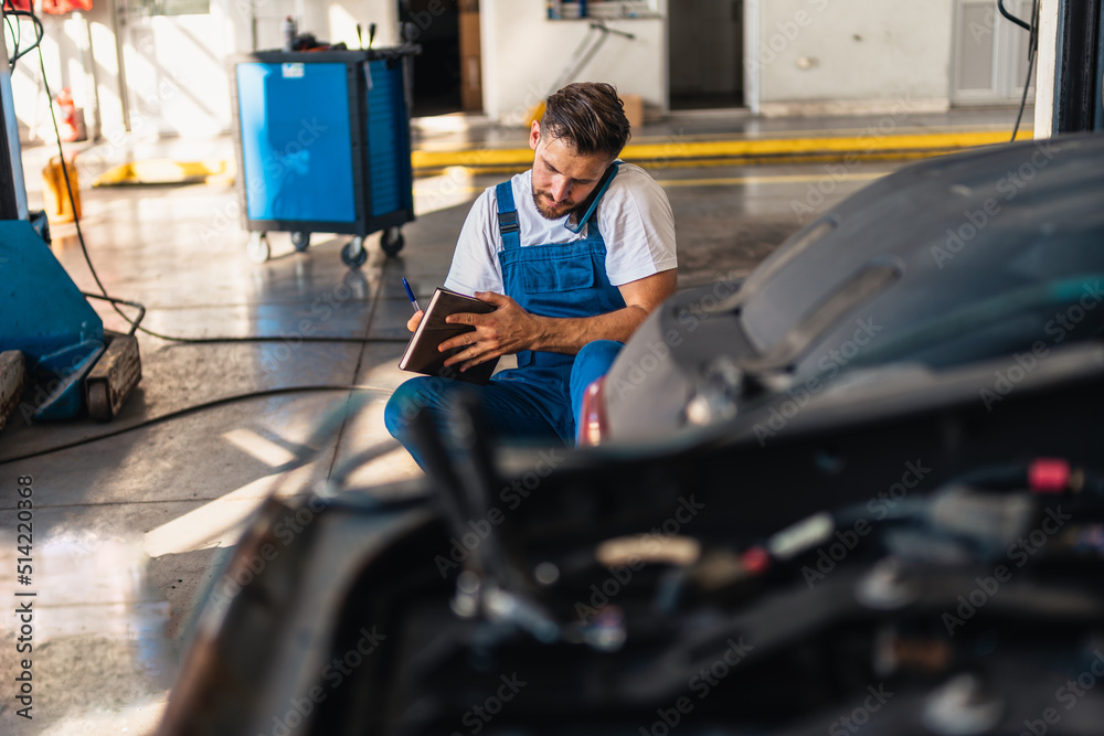 Auto mechanic communicates with client on a smart phone in a car service and writing down what he need to check. Auto mechanic communicates with client on a smart phone in a car service and writing down what he need to check.