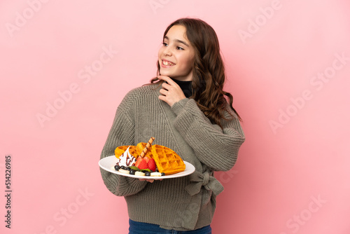 Little girl holding waffles isolated on pink background looking up while smiling