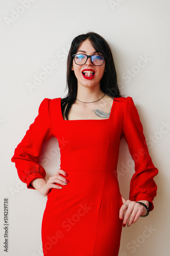 This photograph shows a young attractive brunette woman in a red dress and glasses, chewing gum and smiling.
