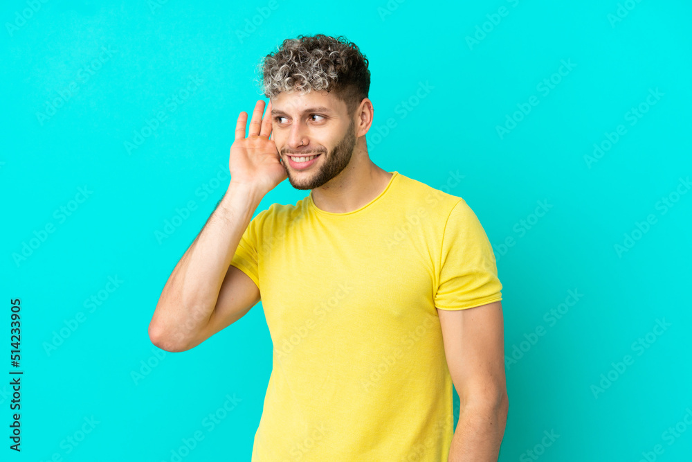 Young handsome caucasian man isolated on blue background listening to something by putting hand on the ear Young handsome caucasian man isolated on blue background listening to something by putting hand on the ear
