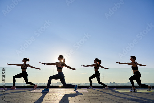 Female silhouettes stand in yoga virabhadrasana warrior pose at group class outdoor on sunrise. Fit healthy young women do yoga fitness exercise meditating together at retreat with sunset summer sky.
