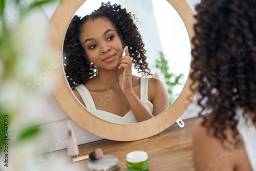 Teen African mixed race girl model touching healthy smooth face skin care looking in bathroom mirror applying facial cream. Teenage beauty skincare morning routine concept.