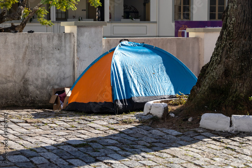 A tent on a city street near a tree. Homeless people in a tent live on the streets of the city. The social problem of unemployment
