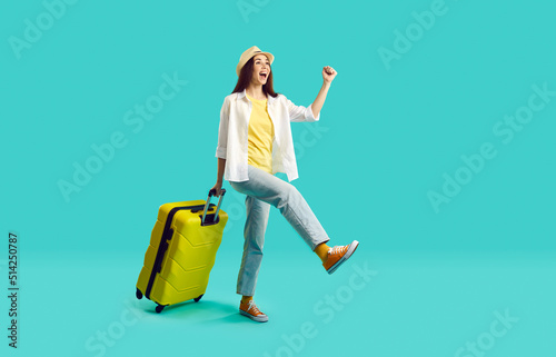 Happy woman with yellow suitcase going on summer holiday. Smiling overjoyed young girl wearing jeans, shirt and panama hat walking with her travel bag on bright turquoise background. Vacation concept