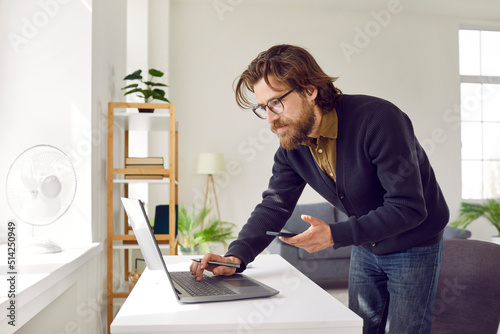 Focused serious smart business man with laptop and mobile phone working from home. Bearded man in casual clothes in home office compares information he sees on screen of laptop and smartphone. © Studio Romantic