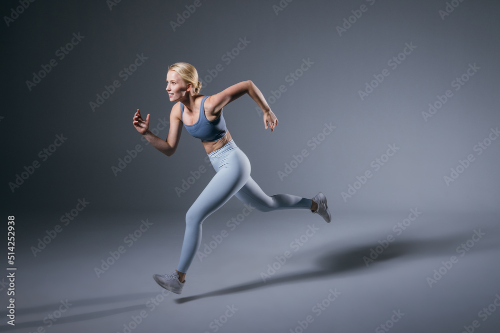 Women running in studio. Female runner on blue gray background. Healthy blonde woman. Profile view of sport runner. Full length of fit girl