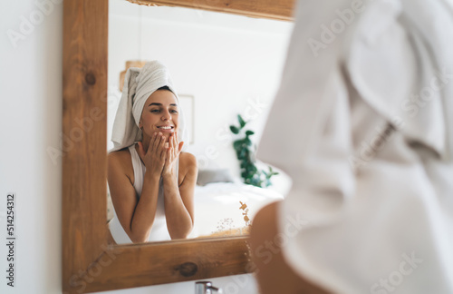 Young beautiful female with towel on head smiling to mirror reflection  standing in the bathroom at home. 30 years old happy woman doing daily morning rituals and cleansing. Enjoying healthy skin care