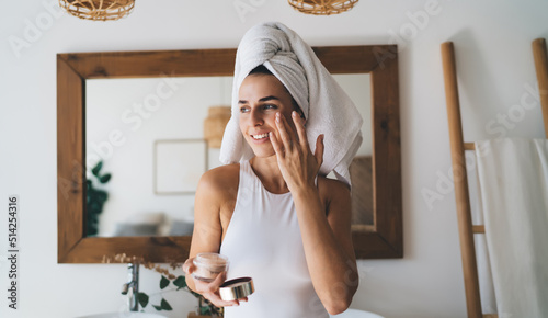 Young smiling female with towel on head puts moisturizing cream on face while standing at home bathroom. 30 years old happy woman doing daily morning rituals. Enjoying healthy skin with collagen care`