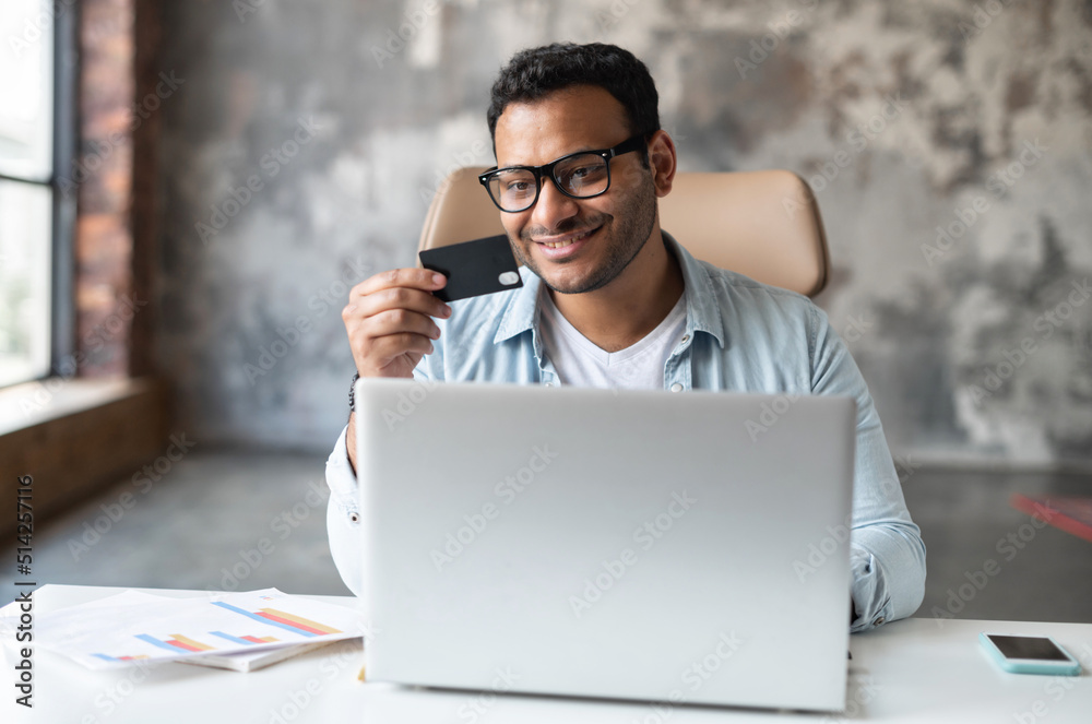 Successful indian businessman in eyeglasses, smiling, sitting at the laptop, paying in the Internet, buying online, working from office,s holding a credit card, buying online, making transaction Successful indian businessman in eyeglasses, smiling, sitting at the laptop, paying in the Internet, buying online, working from office,s holding a credit card, buying online, making transaction