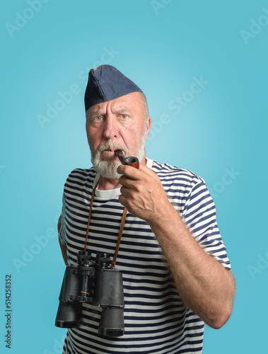 An old sailor smoking a pipe In a marine vest striped shirt and a blue cap - Portrait on light blue © KiNOVO