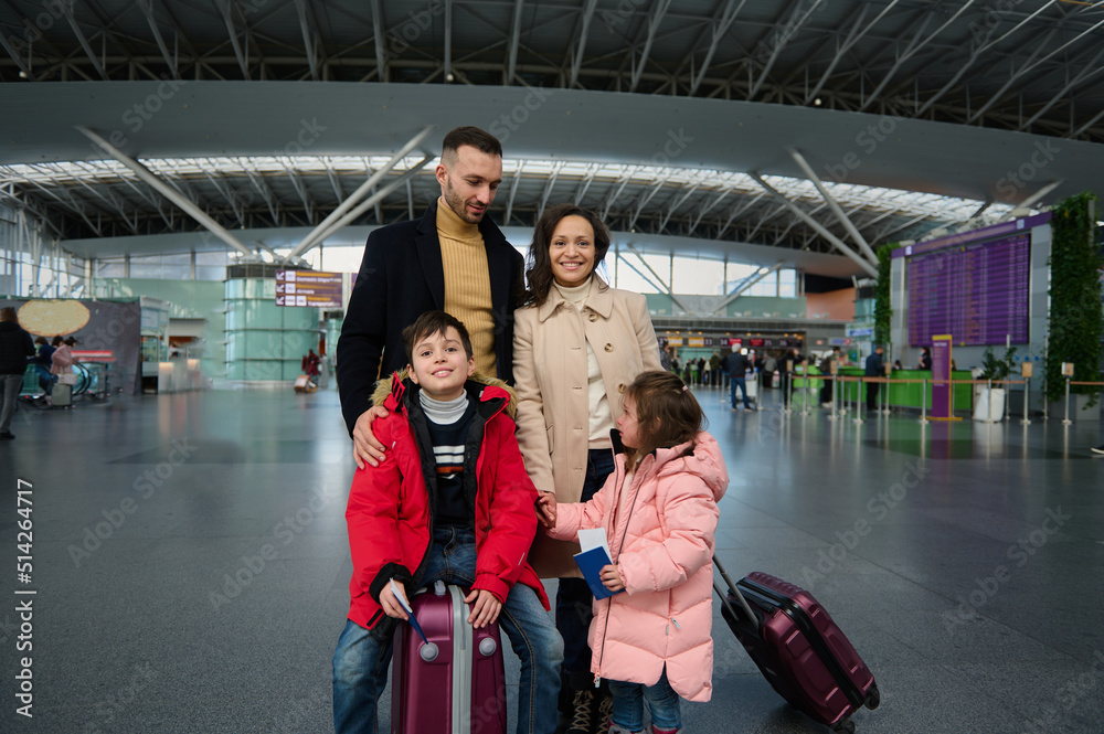 Happy family with kids stand with suitcases in the departure hall of international airport, waiting for flight check-in, customs control and boarding
