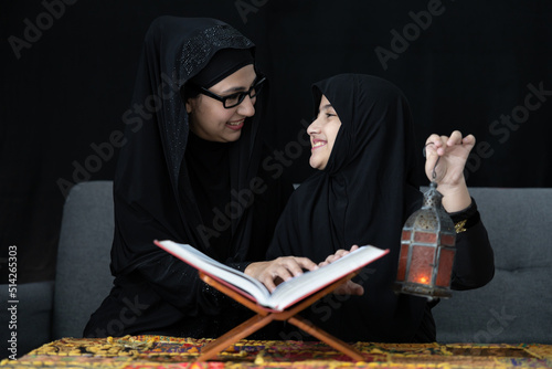 muslim mother with her daughter reading a holy book Quran and holding lamp on black background