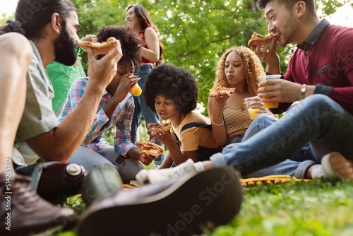 Group of closed friends enjoying dinner in park. Man and women sitting on plaid around pizza and bottles of beer, taking slices from box and eating. Takeaway dinner concept