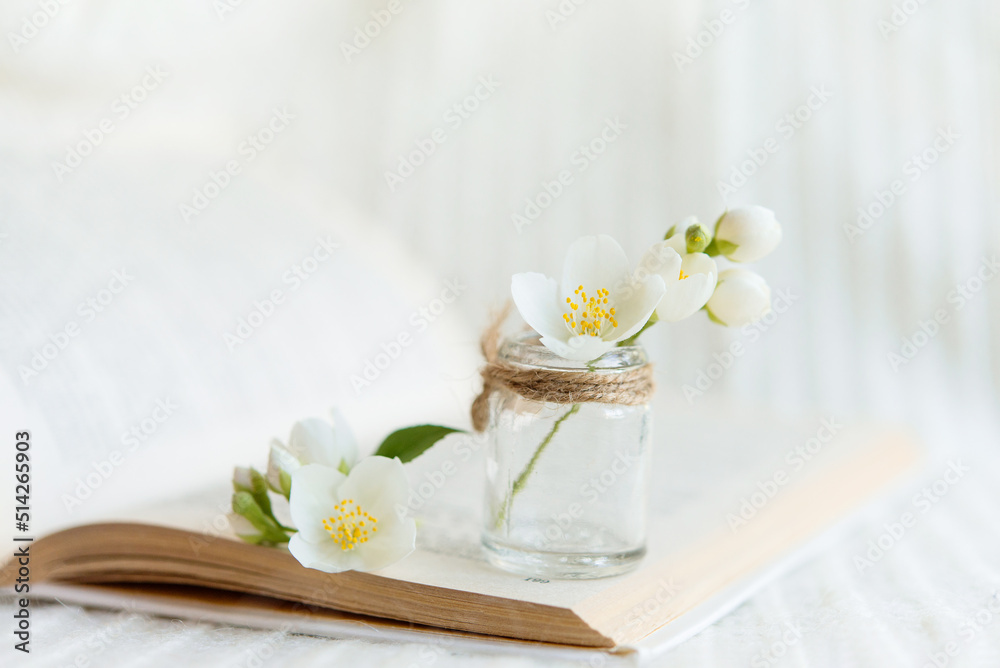 Close-up of a bouquet of white flowers in a blurred glass vase on an open book on a white knitted background. Slow life concept.