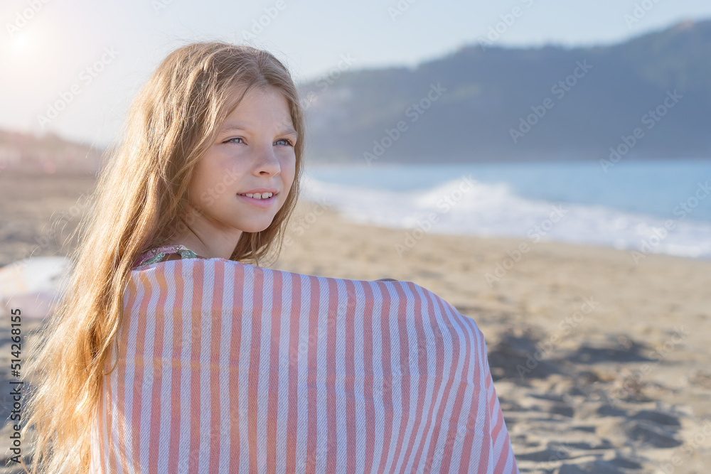 A young girl, covered with a towel, sits on a sandy beach.