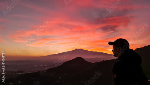 Tourist man watching beautiful sunset behind volcano Mount Etna near Castelmola  Taormina  Sicily  Italy  Europe  EU. Clouds with vibrant red orange colors. Silhouette of person during twilight. Awe