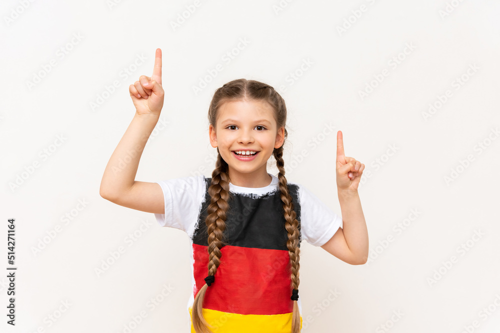 A little girl with a German flag on a T-shirt, with long hair in pigtails, points up with her index finger at your advertisement on a white isolated background. Language courses.