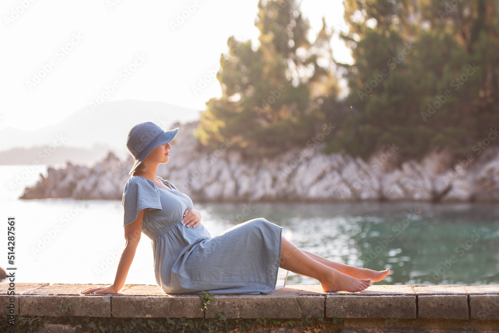 A tender pregnant woman in a denim dress, a blue hat sits by the sea in the sun against the backdrop of a green island. Traveling in pregnancy to Montenegro on the Royal Island. Mothers care and rest